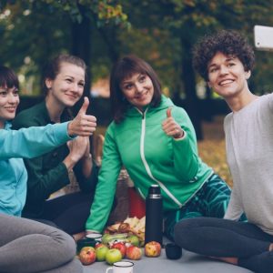 Attractive young ladies yogini are taking selfie using smartphone during picnic in park in autumn. Girls are posing and smiling having fun sitting together on mats.